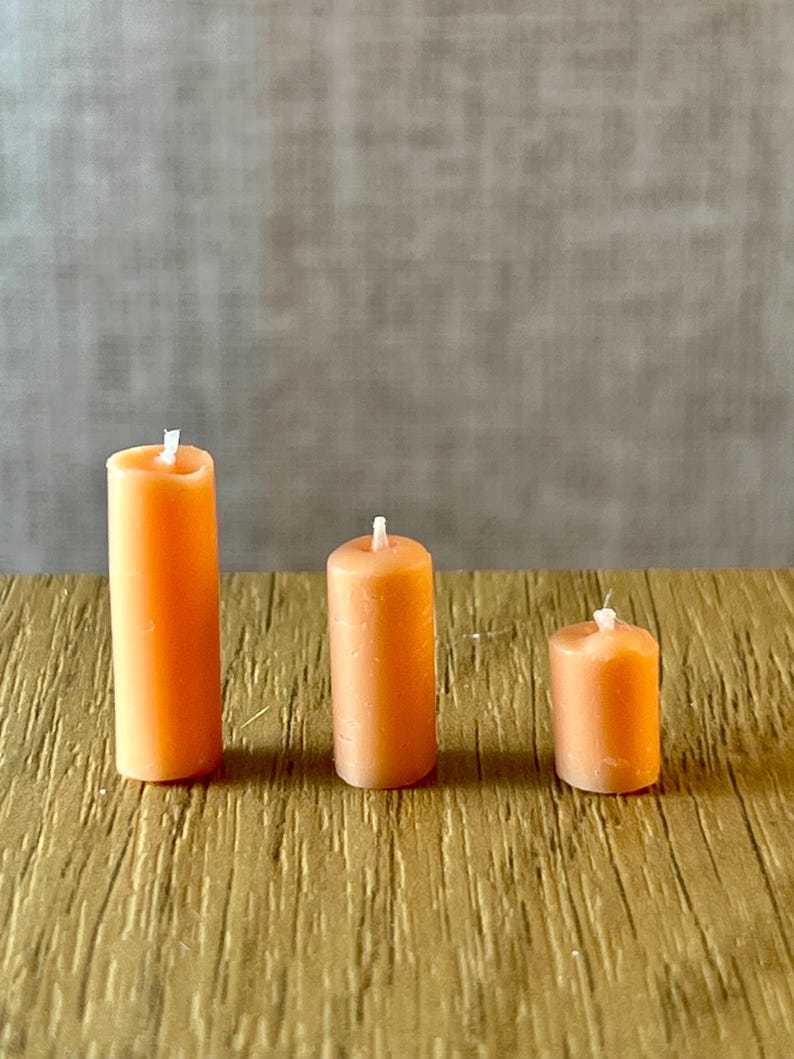 May include: Three orange pillar candles of different heights are displayed on a textured, golden-brown surface. Each candle has a white wick. The background is a neutral grey. The candles are measured in centimetres.