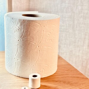 Pode incluir: Um rolo de papel higiênico branco com um padrão floral em relevo. Dois rolos menores de papel higiênico estão na frente do rolo maior. Os itens estão em uma superfície de madeira.