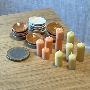 May include: A collection of miniature dollhouse items, including stacks of small brown and white plates, orange and yellow candles, and a Euro coin. The items are arranged on a wooden surface, creating a charming scene.