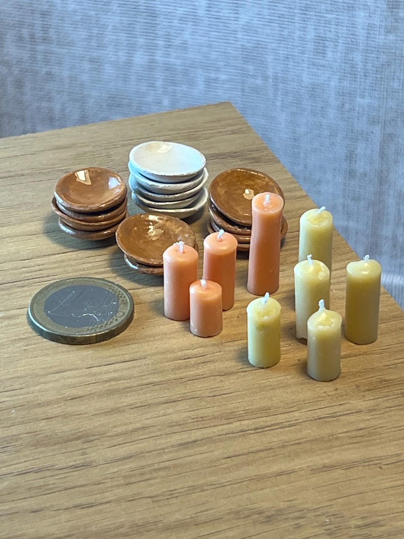 May include: A collection of miniature dollhouse items, including stacks of small brown and white plates, orange and yellow candles, and a Euro coin. The items are arranged on a wooden surface, creating a charming scene.