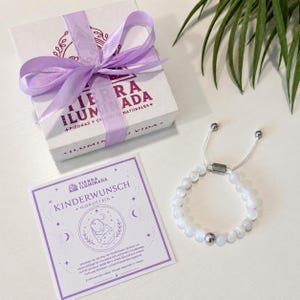 May include: A white gift box with a lavender ribbon, a square card with the text KINDERWUNSCH MONDSTEIN, and a white beaded bracelet with a silver-toned metal bead and an adjustable sliding closure. The box features the text TIERRA ILUMINADA.