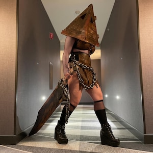 May include: A costume featuring a large, weathered, triangular metal headpiece, a dark brown distressed skirt with a heavy metal chain draped around the waist, black fishnet sleeves, black knee-high socks, and black platform wedge boots.