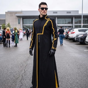 May include: A long black coat featuring a high collar, a front zip, and yellow geometric line patterns along the sleeves and torso. The garment is worn with black sunglasses and black gloves, creating a structured, full-length appearance.
