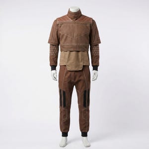May include: A brown two-piece outfit consisting of a short-sleeved top with a high collar, quilted shoulder panels, and white stitching, paired with matching trousers featuring black vertical accents and white seam detailing on a mannequin.