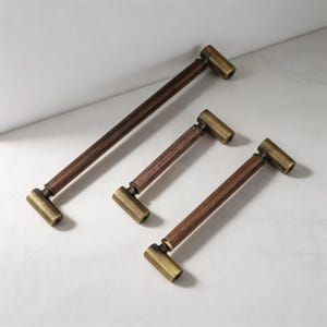 Op de afbeelding: Drie kastgrepen van verschillende lengtes, elk voorzien van een cilindrische donkerbruine houten handgreep verbonden met twee eindkappen van geborsteld messing. De hardware is diagonaal gerangschikt en toont het contrast tussen hout en metaal.