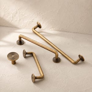 May include: A collection of antique brass hardware including two long drawer pulls with curved ends and circular mounting plates, and one small circular cabinet knob featuring a concentric ring pattern on the face of each piece.