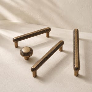 May include: A collection of antique brass hardware including two long rectangular cabinet pulls and one round cabinet knob. Each piece features a textured, knurled surface pattern with a dark, weathered finish and smooth, flat top edges.