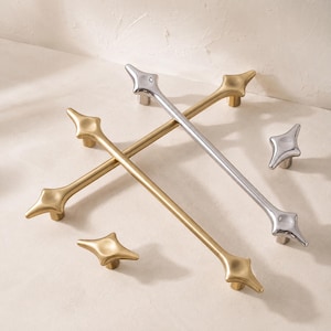 May include: Two long cabinet pulls and two small cabinet knobs. One long pull and one knob have a brushed gold finish, while the other long pull and knob have a polished silver finish. Each piece features a four-pointed star shape at the ends.
