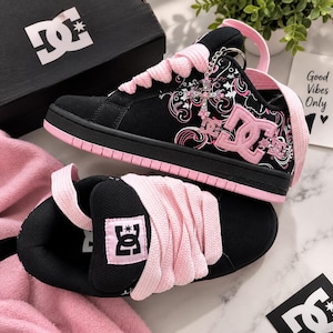 May include: A pair of black and pink DC sneakers featuring thick pink laces, a pink DC logo on the side, and a decorative cross charm attached to the eyelets. The sneakers have a black suede-like upper with pink floral-inspired graphic patterns.