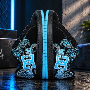 Puede incluir: Un par de zapatillas negras con un logotipo DC azul brillante en el lateral, acentuado con intrincados patrones de remolinos y motivos de estrellas en blanco y azul. Los zapatos tienen cordones negros, parte superior negra y suela azul.