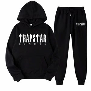 May include: A black hooded sweatshirt and matching sweatpants set. The hoodie features a front kangaroo pocket and the white text TRAPSTAR LONDON printed on the chest. The sweatpants have an elastic waistband with a drawstring and the same white text.
