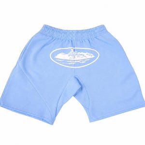 May include: Light blue shorts with an elastic waistband. A white oval graphic with a mountain and cloud design is printed on the back. The shorts are made of a soft material.