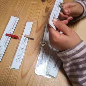 Op de afbeelding: Handen houden een stapel witte flashcards met Franse woorden en zinnen vast. De flashcards worden bij elkaar gehouden met een houten knijper. Andere flashcards met Franse woorden liggen op een houten oppervlak.