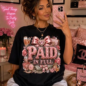 Puede incluir: Una camiseta negra con un estampado gráfico que incluye el texto PAID IN FULL en letras rosas acolchadas. El diseño presenta un gran lazo rosa, copas de champán, un frasco de perfume, un pintalabios, un fajo de billetes, perlas y diamantes.
