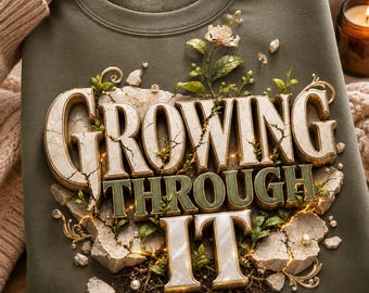 Puede incluir: Una sudadera de cuello redondo color verde oliva con un estampado gráfico del texto "GROWING THROUGH IT" en una fuente 3D con efecto de piedra. Las letras están rodeadas de texturas de piedra agrietada, hojas verdes, enredaderas y perlas.