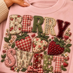 以下が含まれることがあります： 「BERRY SWEET」という文字が大きな布製アップリケで施されたライトピンクのトレーナー。文字と3つの大きなイチゴは、ギンガムチェック、花柄、キルティングなどの様々な柄の生地で作られており、刺繍された花や葉が周囲を飾っています。