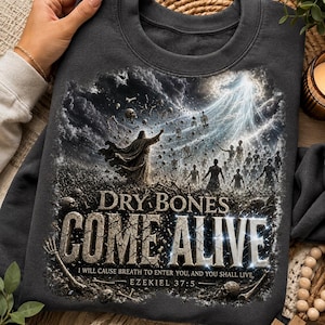 May include: A charcoal gray crewneck sweatshirt featuring a graphic print of a biblical scene with a figure standing before a field of skeletons rising and transforming into people under a bright light, with the text DRY BONES COME ALIVE.