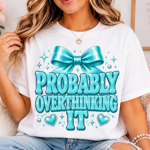 May include: A white t-shirt featuring a large cyan bow graphic at the top, followed by the text "PROBABLY OVERTHINKING IT" in bold cyan letters. The design includes small cyan hearts and sparkling star accents surrounding the text and bow.