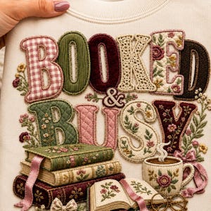 Peut inclure: Un sweat-shirt couleur crème orné de lettres appliquées brodées formant l'inscription BOOKED & BUSY. Le motif comprend des livres empilés, une tasse de café fumante et des détails floraux dans des tons rose, vert et marron.