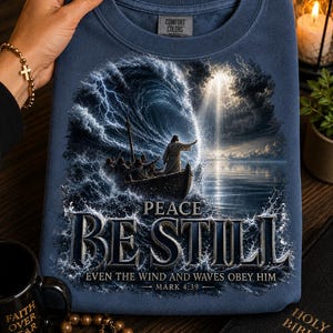 May include: A navy blue crewneck sweatshirt featuring a graphic print of a boat on stormy seas with a figure standing and gesturing toward a large wave. The text reads PEACE BE STILL EVEN THE WIND AND WAVES OBEY HIM MARK 4:39 in metallic lettering.
