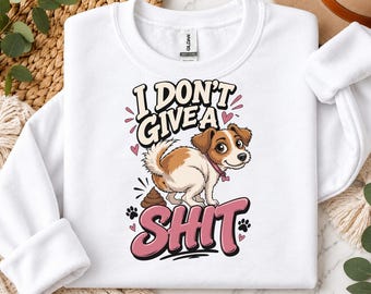 May include: A white crewneck sweatshirt featuring a cartoon illustration of a brown and white dog with a pink collar. The text reads I DON'T GIVE A SHIT in bold lettering, accompanied by small pink hearts and black paw print icons.