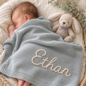 May include: A sleeping baby covered with a pale blue knitted blanket embroidered with the name "Ethan" in cream. A stuffed bunny with a bow tie sits next to the baby in a woven basket.