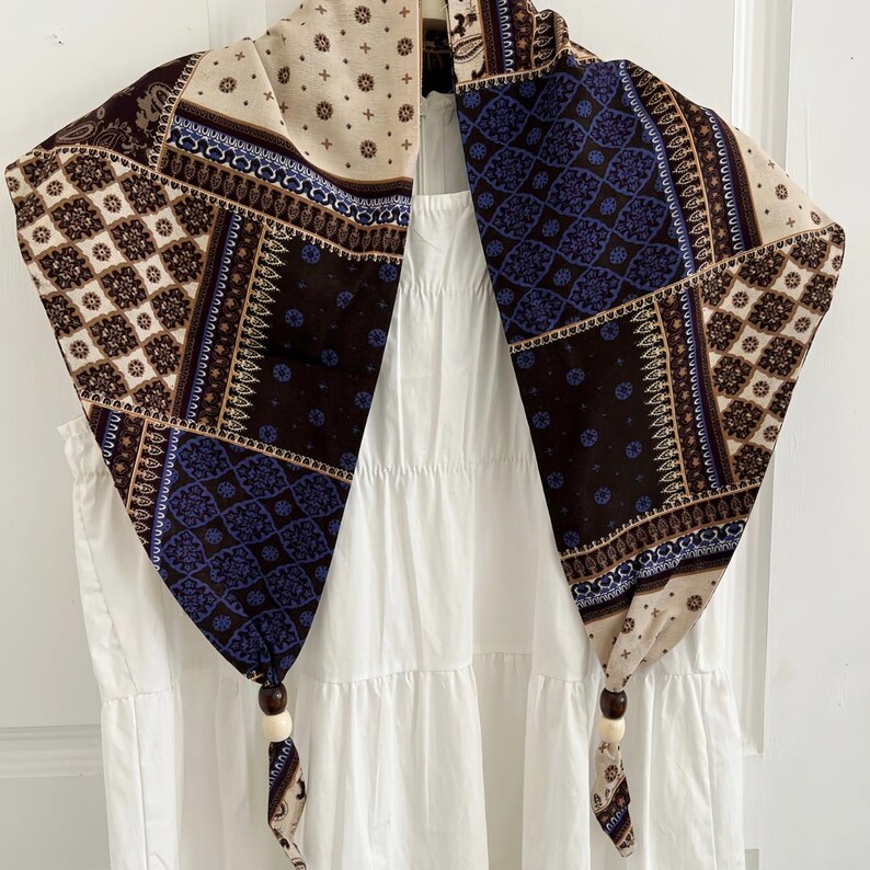 May include: A triangular scarf featuring a patchwork design of cream, dark brown, and deep blue geometric and paisley patterns. The two bottom corners of the scarf are tied with dark brown and cream wooden beads, creating a decorative finish.