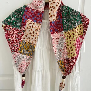 May include: A triangular scarf featuring a patchwork of various floral patterns in red, green, yellow, and white. The scarf is draped over a white tiered dress and finished with two dark brown and cream wooden beads at the bottom corners of the fabric.