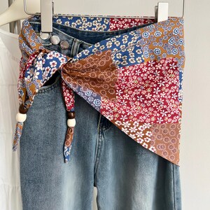 May include: Blue denim jeans featuring a wrap-around fabric belt with a multi-colored floral patchwork pattern. The belt is tied at the side with two dark brown and two cream-colored wooden beads on the ends, secured by silver metal buttons.