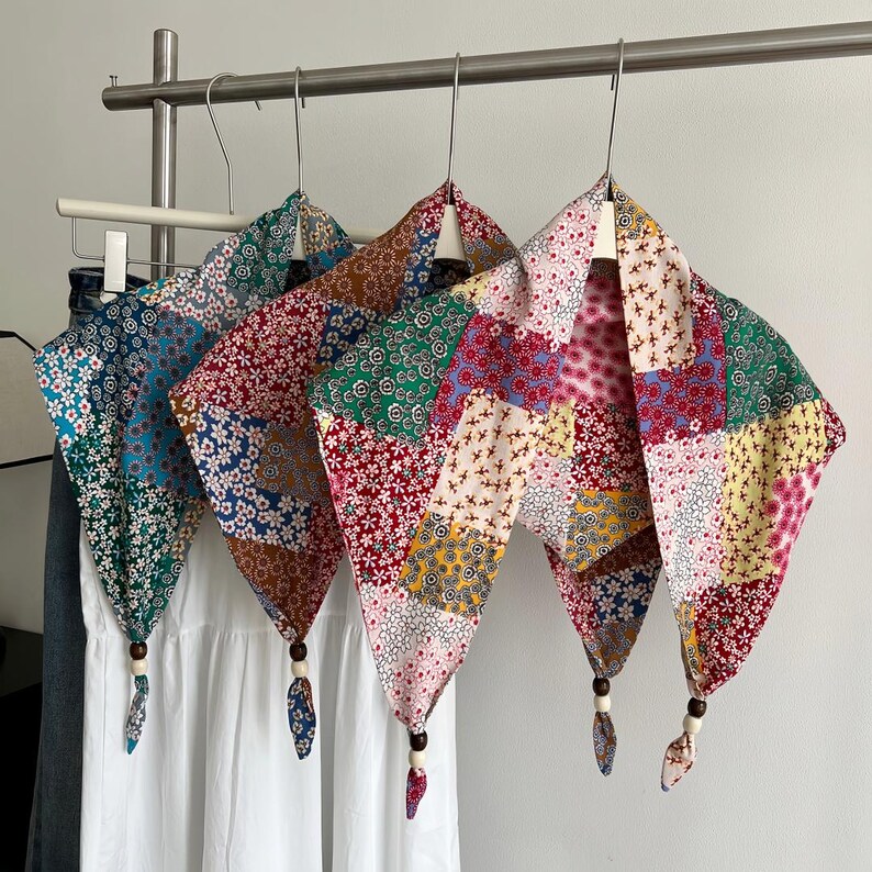 May include: Three triangular scarves featuring a patchwork of various floral patterns in shades of red, blue, green, yellow, and white. Each scarf is finished with a knotted point adorned with a dark brown and a light cream wooden bead.