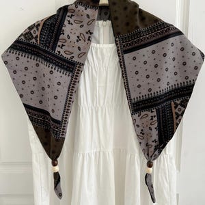 May include: A triangular scarf featuring a patchwork of brown, grey, and black bandana-style patterns with floral and paisley motifs. The two bottom corners are knotted and accented with a dark brown bead and a light cream bead on each side.