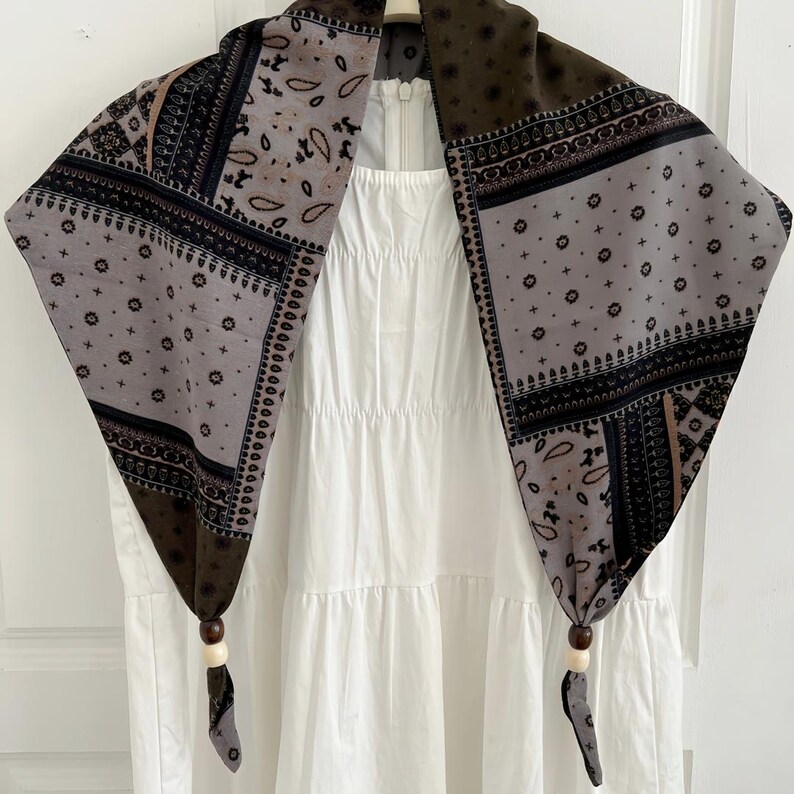 May include: A triangular scarf featuring a patchwork of brown, grey, and black bandana-style patterns with floral and paisley motifs. The two bottom corners are knotted and accented with a dark brown bead and a light cream bead on each side.