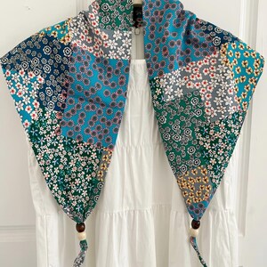 May include: A triangular scarf featuring a patchwork of various floral patterns in shades of teal, navy blue, green, white, and yellow. The two bottom corners of the scarf are finished with dark brown and cream-colored wooden beads on fabric ties.