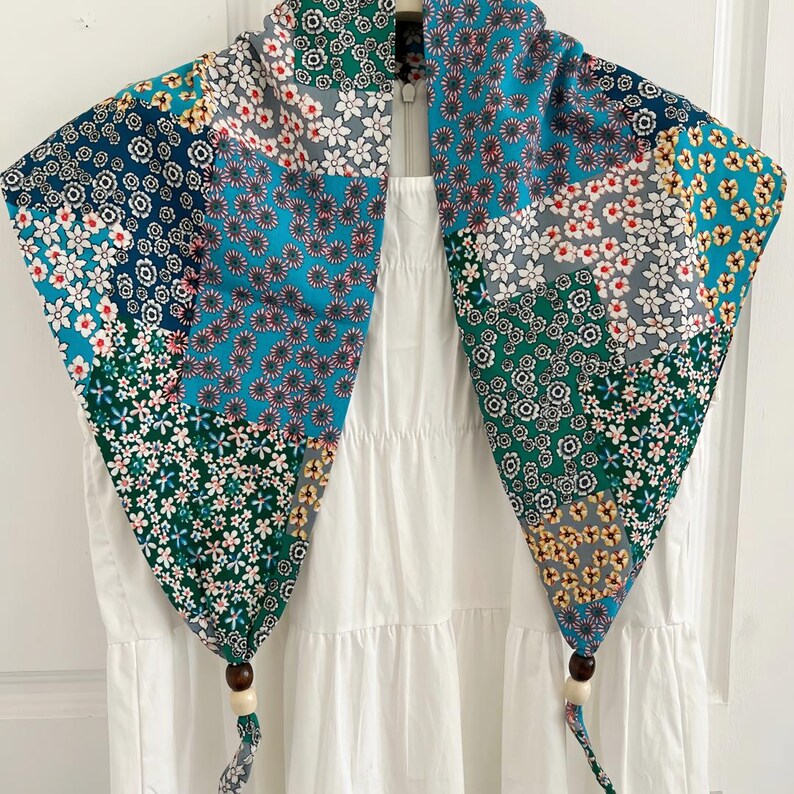 May include: A triangular scarf featuring a patchwork of various floral patterns in shades of teal, navy blue, green, white, and yellow. The two bottom corners of the scarf are finished with dark brown and cream-colored wooden beads on fabric ties.
