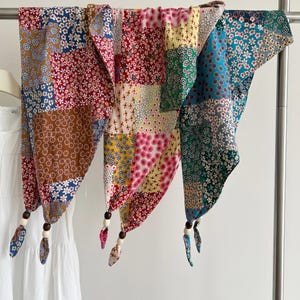 May include: A patchwork scarf featuring a variety of floral patterns in red, blue, yellow, green, and white. The scarf is constructed from multiple fabric squares and includes small fabric tails at the bottom corners, each accented with wooden beads.