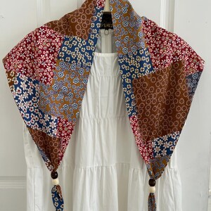 May include: A white sleeveless dress with a tiered skirt and a zipper closure at the back, paired with a triangular scarf featuring a patchwork of floral patterns in shades of red, blue, and brown, finished with dark and light wooden beads.