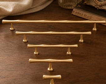 May include: A collection of five gold-toned metal cabinet pulls and knobs arranged in a vertical row by size. Each piece features a smooth, cylindrical bamboo-inspired design with segmented nodes and flared mounting posts for attachment to furniture.