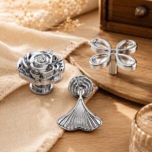 May include: Three silver-toned metal cabinet knobs with distinct shapes. One knob is shaped like a blooming rose, another features a four-petaled clover design, and the third is a fan-shaped pull with a circular base and vertical ribbed detailing.