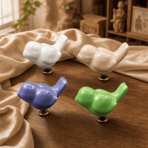 May include: Four ceramic bird-shaped cabinet knobs featuring a glossy finish in white, beige, purple, and bright green. Each bird figure is mounted on a small, circular, dark metallic base, designed for attachment to furniture drawers or doors.