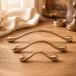 May include: Three curved cabinet pulls in a brushed brass finish, arranged in a descending size order from top to bottom. Each handle features a smooth, arched design with rounded, teardrop-shaped ends, showcasing a consistent metallic texture.