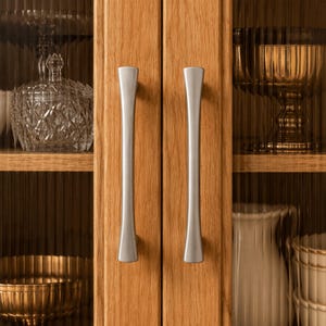 May include: Two vertical brushed silver metal cabinet handles mounted on light brown oak wood cabinet doors. Behind the glass panels, a clear crystal bowl, a gold-toned fluted pedestal dish, and a white ceramic pitcher are visible.
