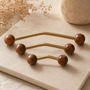 May include: Three cabinet pulls of varying lengths, each featuring a brushed gold metal bar with a slight centre angle and a smooth, round dark brown wooden knob at each end, arranged in a descending size order from back to front.