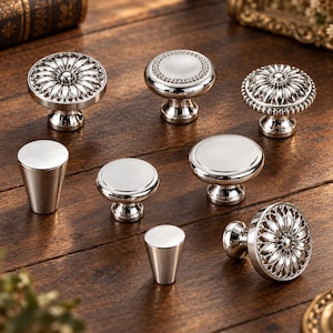 May include: A collection of eight silver-toned metal cabinet knobs in various shapes and designs. The set includes round knobs with intricate floral petal patterns, smooth domed surfaces, beaded edges, and two tapered cylindrical knobs with flat tops.
