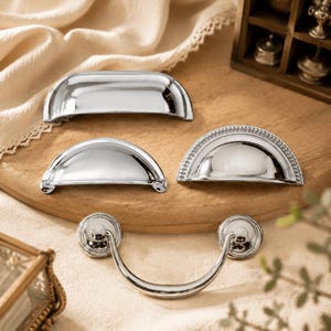 May include: A collection of four silver-toned metal drawer pulls. Three are semi-circular cup pulls, with one featuring a beaded edge detail. The fourth is a drop-style pull with a curved handle attached to two circular mounting plates.