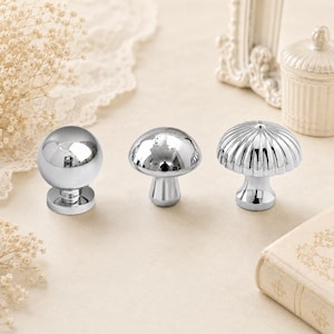 May include: Three silver-toned metal cabinet knobs with a reflective finish. The first knob is a smooth sphere, the second is a mushroom-shaped dome, and the third features a fluted, ribbed design on the top surface with a cylindrical base.