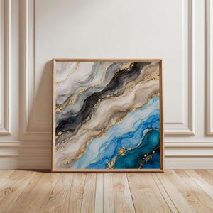 Puede incluir: Una pieza de arte mural cuadrada en un marco de madera clara con una pintura fluida abstracta. La composición muestra ondas fluidas y superpuestas de negro, gris, beige y azul vibrante, acentuadas con finas líneas doradas.