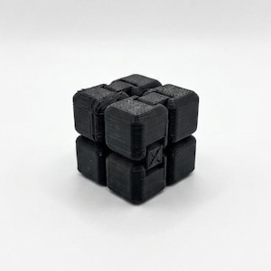 Può includere: Un cubo nero stampato in 3D con un design complesso. Il cubo è composto da blocchi più piccoli e strutturati e presenta un dettaglio unico a forma di X su un lato. L'oggetto è posto su uno sfondo bianco.
