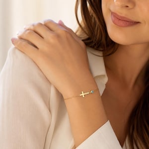 May include: A delicate gold chain bracelet featuring a small horizontal cross charm and a single round blue gemstone accent. The bracelet is crafted with a fine link chain and a secure clasp, presenting a minimalist design that rests on the wrist.