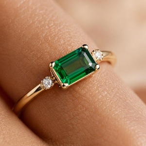 May include: A gold ring featuring a central emerald-cut green gemstone flanked by two small round white diamonds. The band is polished yellow gold, and the setting holds the stones in a horizontal orientation on the finger.