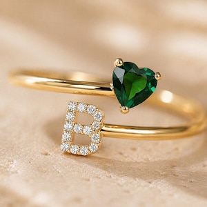 Op de afbeelding: Een gouden bypass-ring met een hartvormige donkergroene edelsteen aan het ene uiteinde en een hoofdletter B ingelegd met kleine heldere ronde steentjes aan het andere uiteinde. De ring is van gepolijst goud en heeft een open ontwerp.